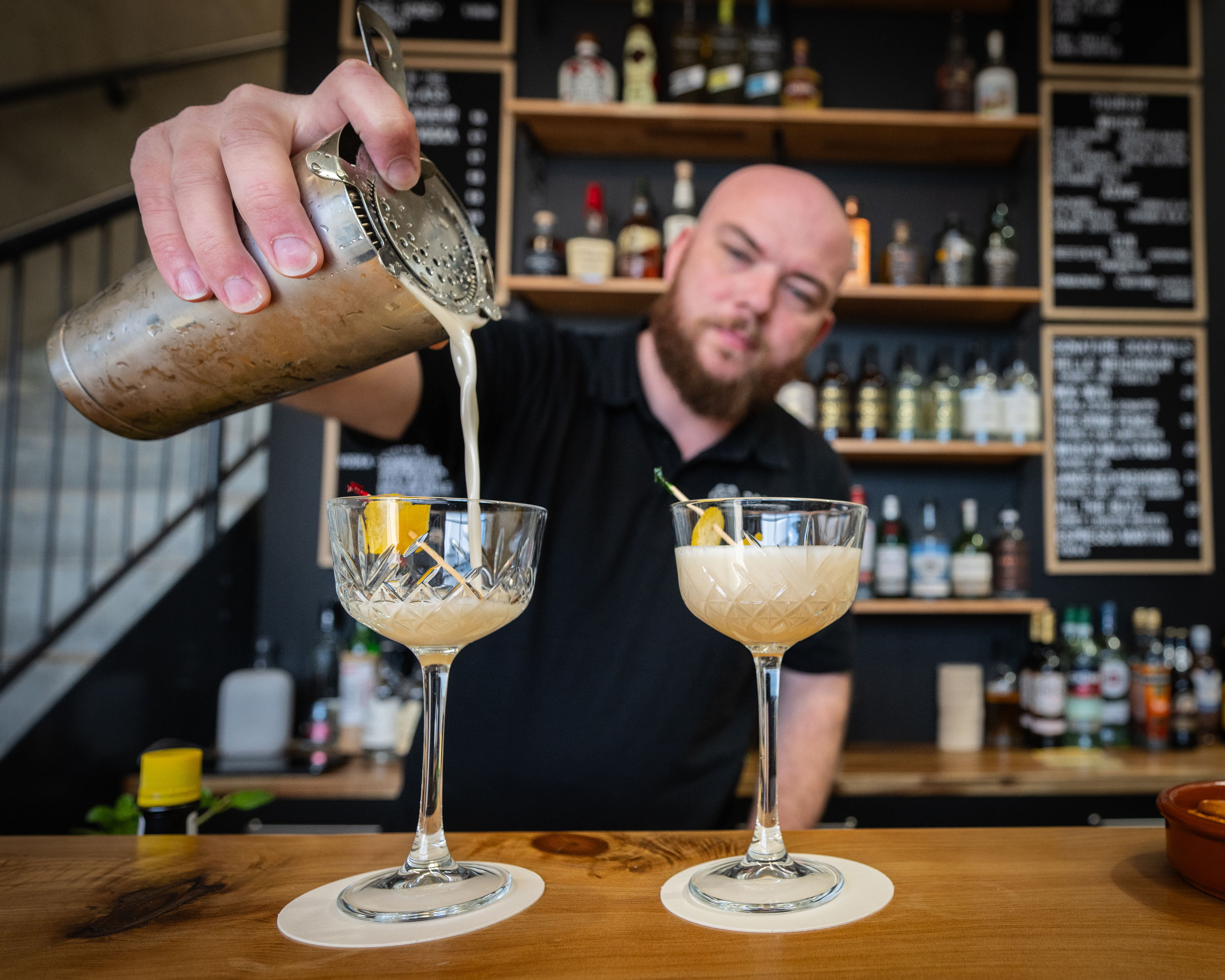 A man pouring a drink into two glasses with a drink setup in the background